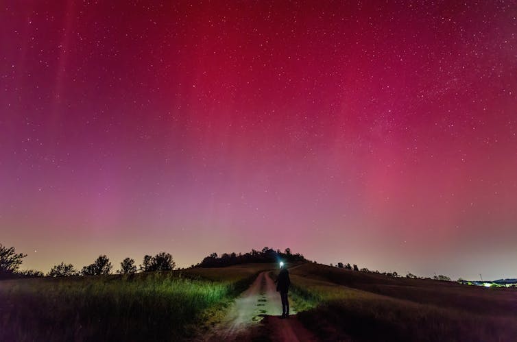 A person standing on a dark road at night looking up at a bright pink-red sky.